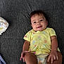 Sage joined the competition — help win amazing prizes! baby, infant, smiling, yellow_shirt, diaper, lying_down, dark_background, happy, cute, child, person, toddler, skin, face, hand, foot, clothing, indoors, texture, playful
