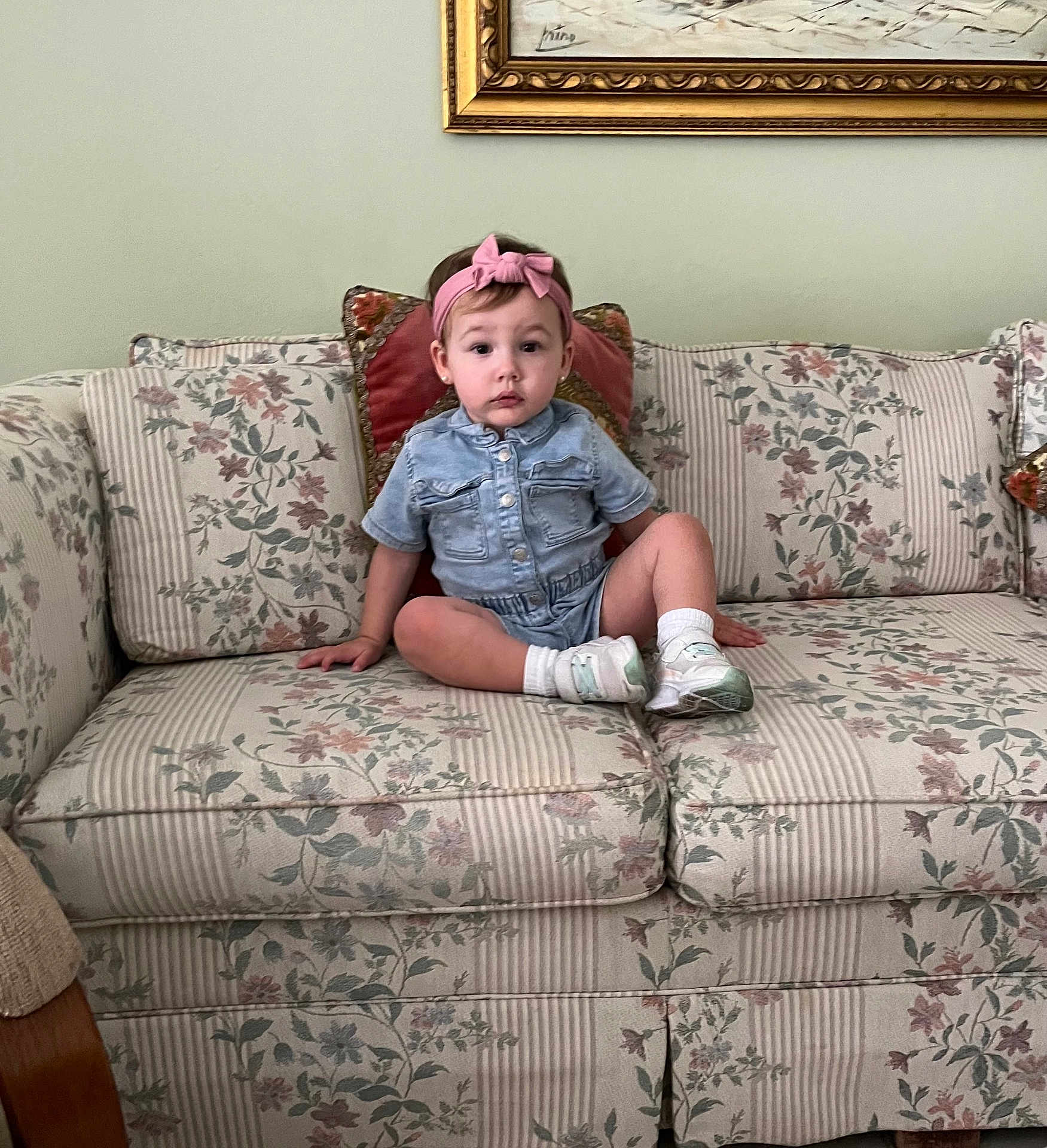 Ellie is registered to the contest to win money with this photo: toddler, child, denim, romper, pink_headband, sitting, couch, floral_pattern, cushions, indoor, living_room, painting, wall_art, footwear, socks, curious, neutral_expression, furniture, person, portrait