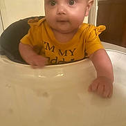 Gwendolyn is registered to the contest to win money with this photo: baby, infant, child, high_chair, yellow_shirt, face, eyes, hand, tray, closeup, portrait, indoor, dining_room, soft_focus, curious, sitting, cute, chubby_cheeks, furniture, gaze