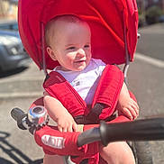 Elias is registered to the contest to win money with this photo: baby, child, tricycle, red_seat, outdoor, sunny, daytime, smile, helmet_absent, safety_harness, urban, sidewalk, street, handlebar, bell, clothing, person, infant, seat, blue_sky