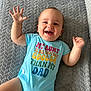 baby, child, smiling, onesie, blue_clothing, text_on_clothing, hand_raised, happy, lying_down, blanket, indoor, infant, cute, portrait, small_child, cheerful, person, face, skin, casual