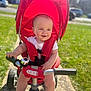 baby, child, tricycle, stroller, red, outdoor, sunlight, grass, smile, happy, person, handlebar, foot, toy, pathway, daylight, park, vehicle, infant, seat