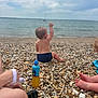 toddler, child, beach, water, sea, pebbles, sitting, back_view, drink_bottle, sky, cloudy, person, foot, hand, shore, outdoor, relaxation, summer, nature, casual