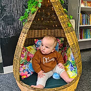 Atlas is registered to the contest to win money with this photo: baby, child, wicker_chair, indoor, cozy, sweater, floral_cushions, leafy_vines, bookshelf, carpet, curious, seated, pod_chair, decor, soft_lighting, floor, infant, casual_clothing, indoors, portrait