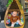 baby, child, wicker_chair, indoor, cozy, sweater, floral_cushions, leafy_vines, bookshelf, carpet, curious, seated, pod_chair, decor, soft_lighting, floor, infant, casual_clothing, indoors, portrait