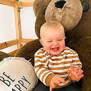 Nolan participe au concours pour gagner de l'argent avec cette photo : baby, child, smiling, drool, plush_bear, striped_sweater, pillow, text_on_pillow, wooden_frame, ivy, indoor, happy, cute, sitting, clothing, person, face, hand, toddler, cozy