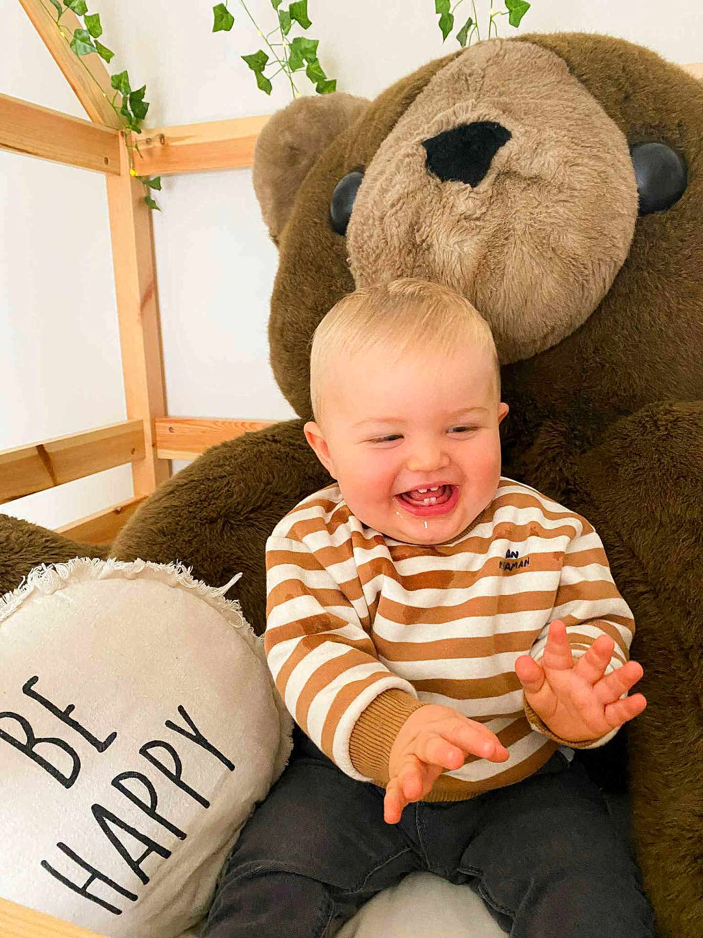 Nolan participe au concours pour gagner de l'argent avec cette photo : baby, child, smiling, drool, plush_bear, striped_sweater, pillow, text_on_pillow, wooden_frame, ivy, indoor, happy, cute, sitting, clothing, person, face, hand, toddler, cozy