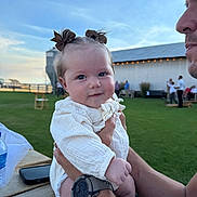Adalyn joined the competition — help win amazing prizes! baby, child, person, adult, holding, outdoor, grass, barn, sky, watch, table, water_bottle, phone, bow, white_clothing, smile, face, hand, people, daylight