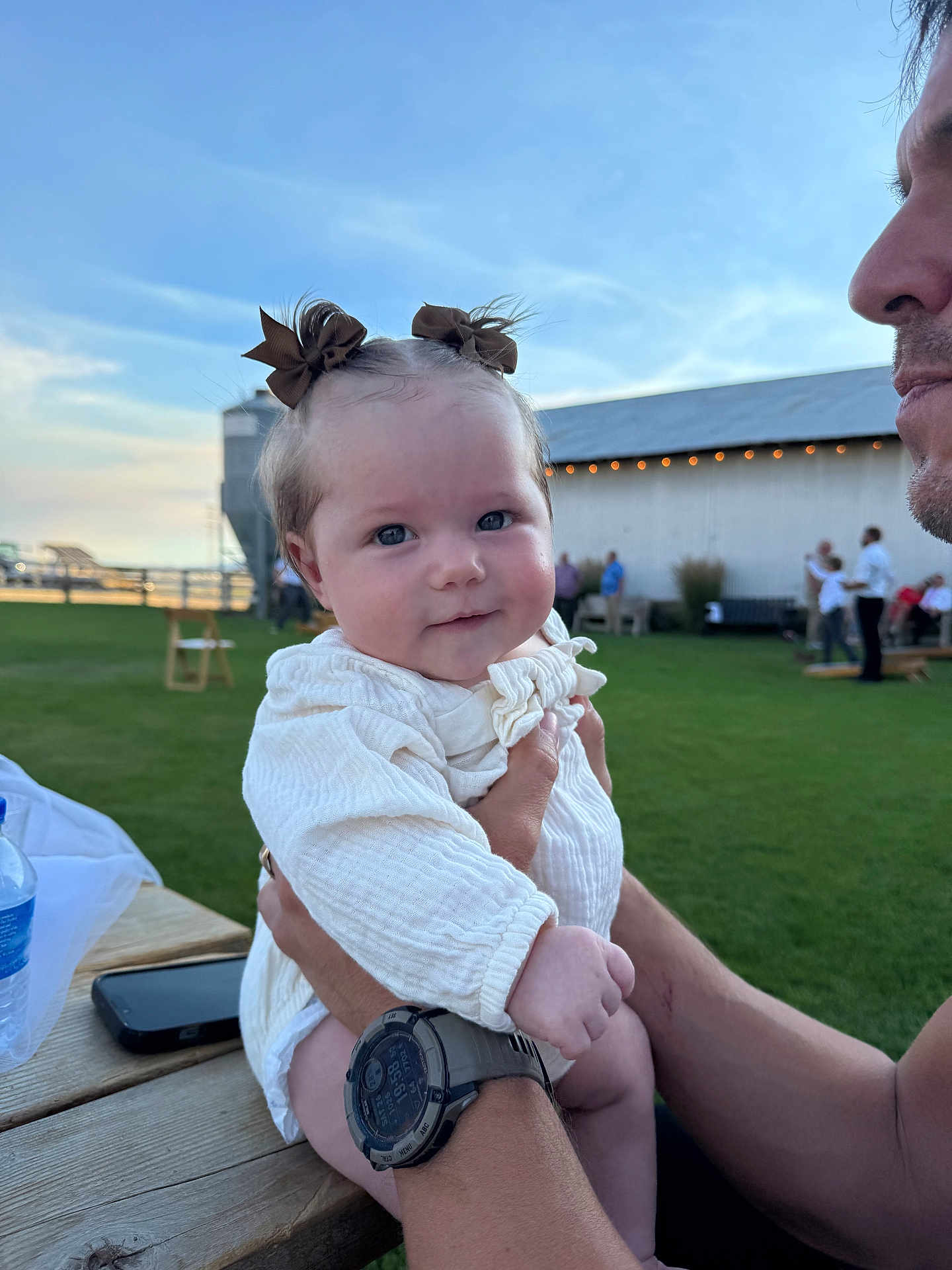 Adalyn joined the competition — help win amazing prizes! baby, child, person, adult, holding, outdoor, grass, barn, sky, watch, table, water_bottle, phone, bow, white_clothing, smile, face, hand, people, daylight