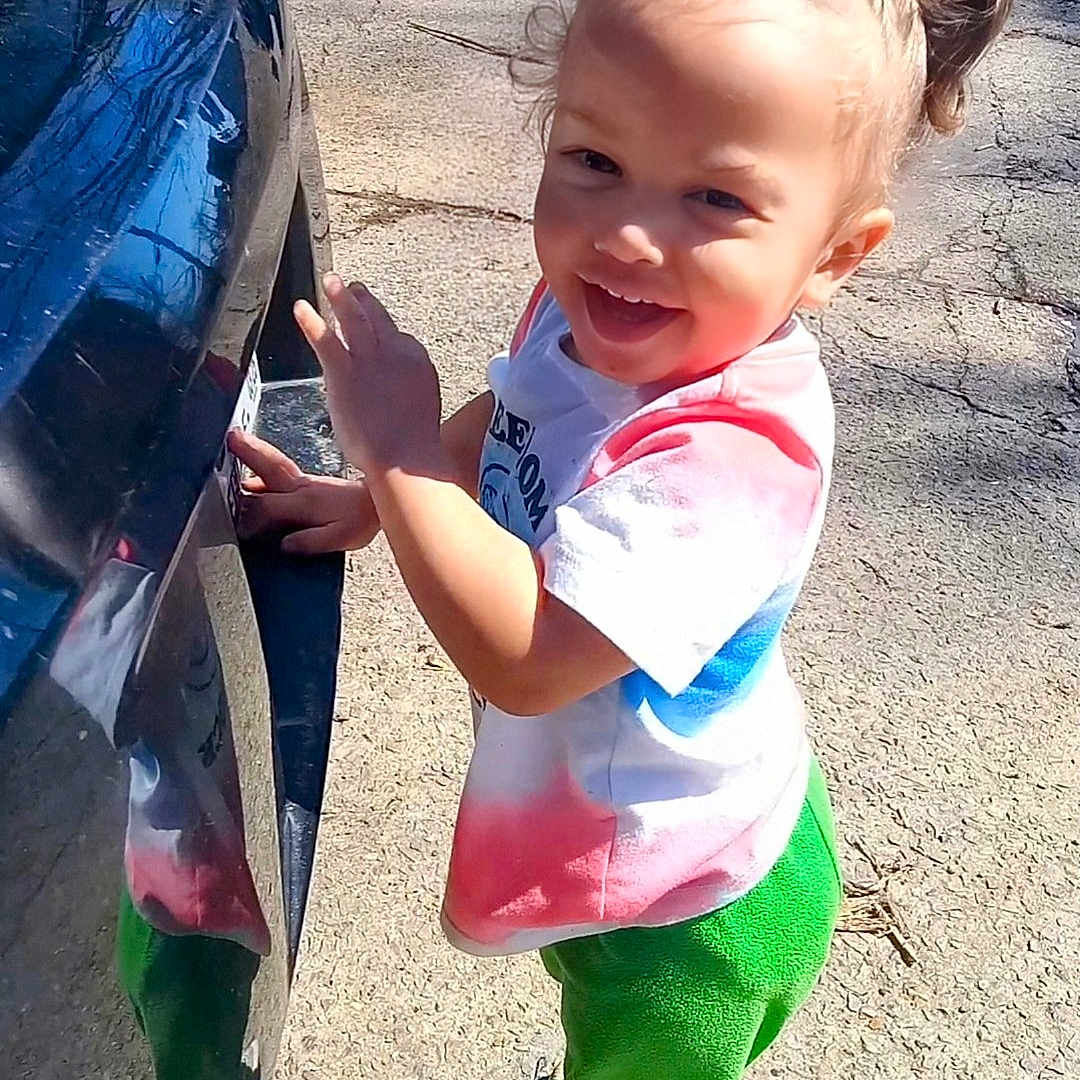 River joined the competition — help win amazing prizes! braided_hair, car, casual_clothing, child, cute, daylight, double_buns, green_pants, happy, outdoor, pavement, person, playful, reflection, smiling, sneakers, standing, sunlight, tie_dye_shirt, toddler