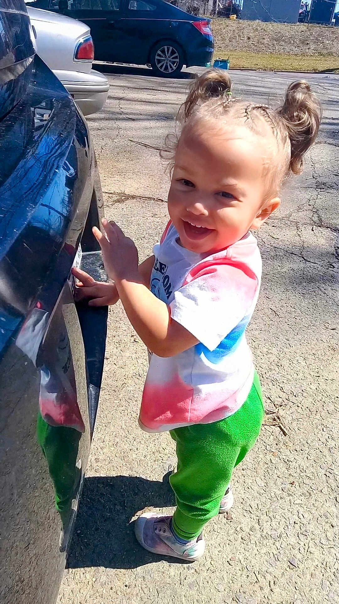 River joined the competition — help win amazing prizes! toddler, child, smiling, standing, outdoor, car, braided_hair, double_buns, tie_dye_shirt, green_pants, sneakers, sunlight, pavement, reflection, happy, playful, person, daylight, cute, casual_clothing