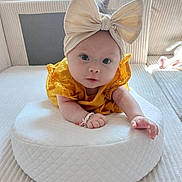 Mya a rejoint le concours — aidez-le/la à gagner de superbes lots ! baby, infant, headband, bow, yellow_dress, cushion, white_cushion, soft_toys, indoor, face, hands, curious, child, portrait, cozy, home, cute, light, soft_texture, comfortable