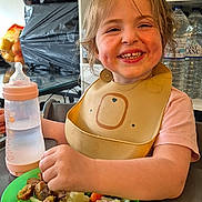 Kaylie participe au concours pour gagner de l'argent avec cette photo : child, toddler, smiling, baby_bottle, high_chair, food, vegetables, plate, bib, kitchen, shelf, cartons, water_bottle, person, meal, table, happy, indoors, face, eating