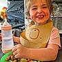 child, toddler, smiling, baby_bottle, high_chair, food, vegetables, plate, bib, kitchen, shelf, cartons, water_bottle, person, meal, table, happy, indoors, face, eating