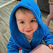 Lyam participe au concours pour gagner de l'argent avec cette photo : toddler, child, blue_robe, hooded_clothing, smile, indoor, floor, hand, face, eyes, person, baby, cute, wooden_floor, background, furniture, shelf, toy, home, playful