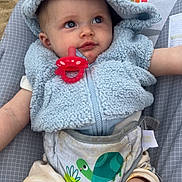 Mateo is registered to the contest to win money with this photo: animal_print, baby, baby_seat, blue_hoodie, child, curious, cute, hood, infant, legs, outdoor, pacifier, pattern, plush_clothing, sand, seatbelt, sitting, skin, toddler, turtle_print