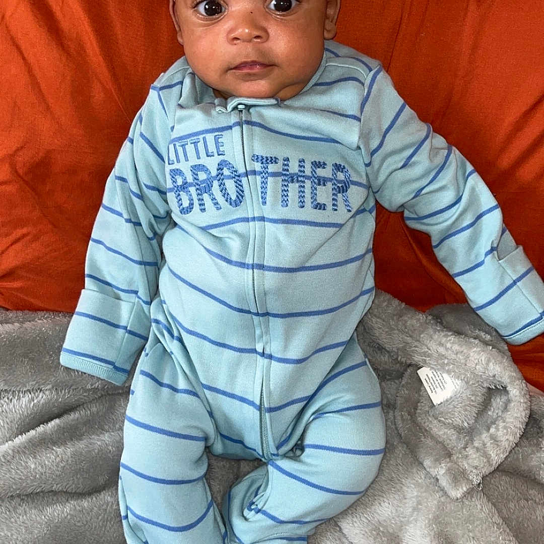 Jakai Kyrie is registered to the contest to win money with this photo: baby, blanket, blue, child, cozy, cute, face, indoor, infant, lying_down, onesie, orange, person, pillow, portrait, sleepwear, soft_texture, striped_clothing, wide_eyes, young