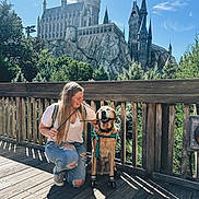 Jake is registered to the contest to win money with this photo: woman, dog, leash, harness, wand, castle, theme_park, bridge, wooden_deck, railing, trees, sunlight, shadow, smile, jeans, kneeling, rocky_cliff, tourist, dog_boots, outdoor