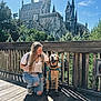 woman, dog, leash, harness, wand, castle, theme_park, bridge, wooden_deck, railing, trees, sunlight, shadow, smile, jeans, kneeling, rocky_cliff, tourist, dog_boots, outdoor