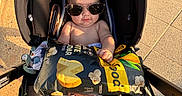 Aryes is registered to the contest to win money with this photo: baby, infant, child, stroller, sunglasses, snack_bag, food, popcorn, outdoor, sunlight, pavement, tiles, portrait, sitting, 裸, feet, blanket, car_seat, fashion, funny