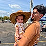 baby, child, adult, cowboy_hat, sunglasses, carrying, portrait, outdoor, sunny, parking_lot, pavement, sky, clouds, casual_clothing, onesie, sneakers, smiling, family, street_scene, photography