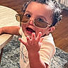 child, toddler, curly_hair, sunglasses, smiling, open_mouth, hand, reaching, portrait, close_up, indoor, home, table, wooden_floor, rug, casual_clothing, white_shirt, fashion, happy, candid