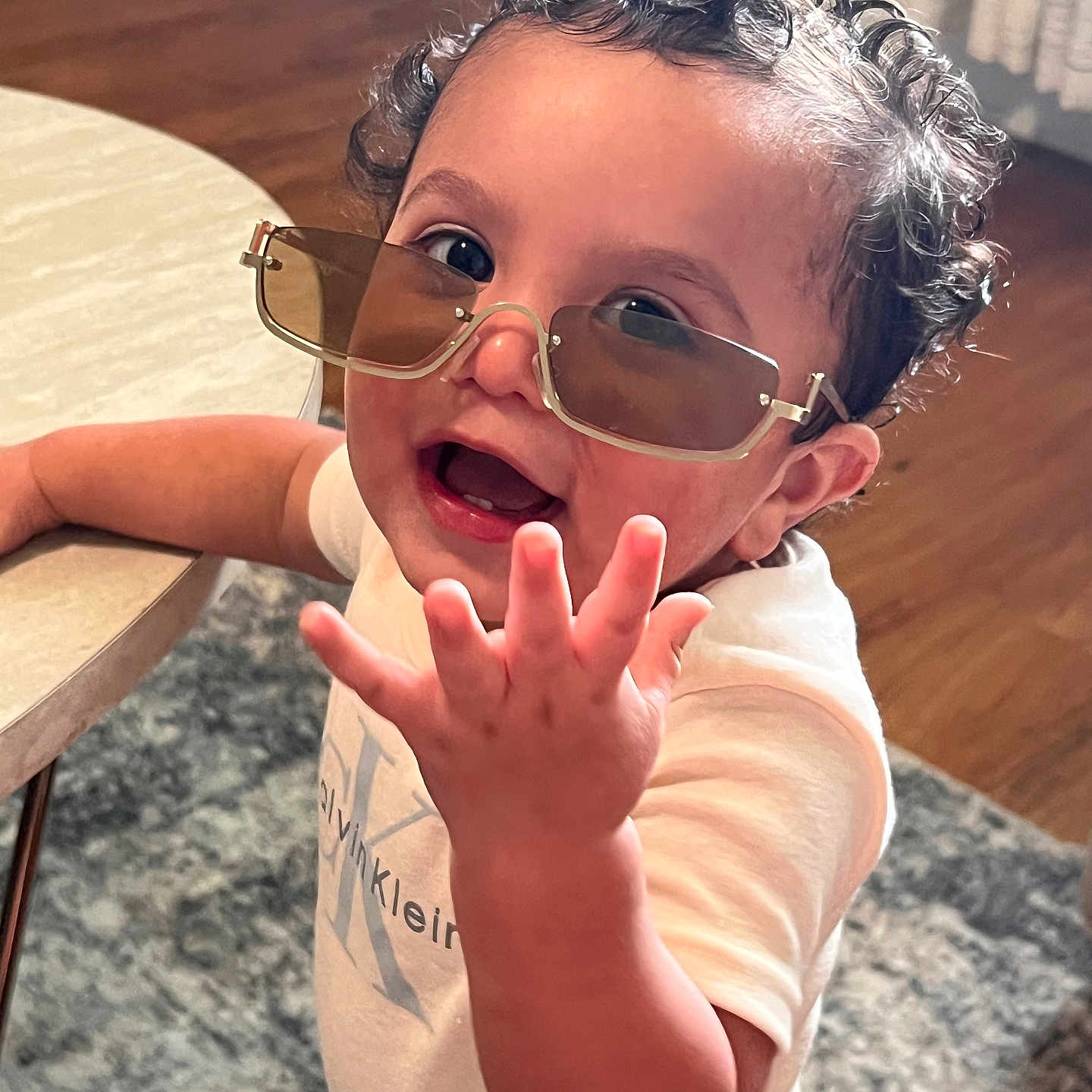 Rowen joined the competition — help win amazing prizes! child, toddler, curly_hair, sunglasses, smiling, open_mouth, hand, reaching, portrait, close_up, indoor, home, table, wooden_floor, rug, casual_clothing, white_shirt, fashion, happy, candid