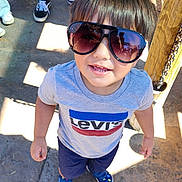 Dominic is registered to the contest to win money with this photo: child, toddler, sunglasses, smiling, outdoor, playground, portrait, fashion, casual_clothing, levis_shirt, shorts, blue_shoes, wooden_post, chain, sunlight, shadows, pavement, people, close_up, happy