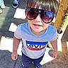 child, toddler, sunglasses, smiling, outdoor, playground, portrait, fashion, casual_clothing, levis_shirt, shorts, blue_shoes, wooden_post, chain, sunlight, shadows, pavement, people, close_up, happy