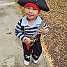 child, toddler, pirate, pirate_costume, hat, skull_and_crossbones, toy_sword, striped_shirt, costume, outdoor, sidewalk, grass, leaves, portrait, standing, smiling, playful, full_body, daylight, photography