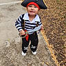 child, toddler, pirate, costume, hat, toy_sword, skull_pattern, striped_shirt, outdoor, sidewalk, residential_area, smiling, standing, portrait, full_body, playtime, dress_up, tree, grass, photography