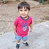 child, boy, toddler, portrait, smiling, standing, outdoor, courtyard, pavement, pink_shirt, peace_sign, shorts, sneakers, socks, dark_hair, brown_hair, face, full_body, daylight, plants