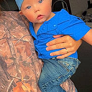 Jerry is registered to the contest to win money with this photo: baby, infant, child, blue_hat, blue_shirt, jeans, bare_foot, blue_eyes, hand, ring, camo_blanket, holding, portrait, indoor, close_up, cap_backwards, curious_expression, cute, denim_pants, person