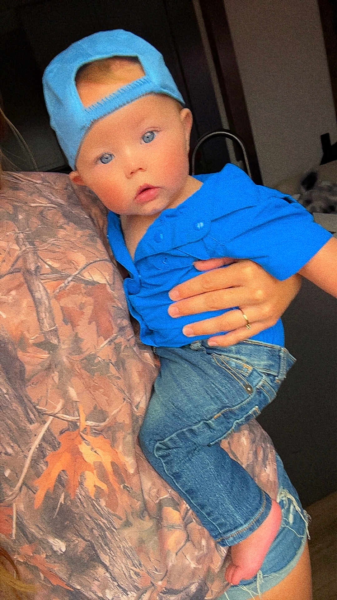 Jerry is registered to the contest to win money with this photo: baby, infant, child, blue_hat, blue_shirt, jeans, bare_foot, blue_eyes, hand, ring, camo_blanket, holding, portrait, indoor, close_up, cap_backwards, curious_expression, cute, denim_pants, person