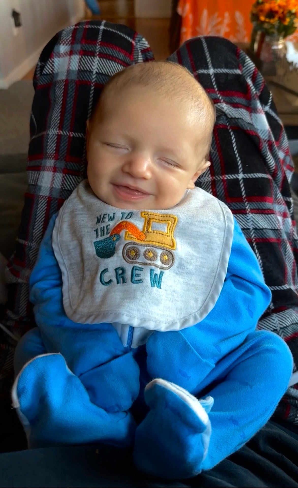 Grayson joined the competition — help win amazing prizes! baby, smiling, blue_onesie, bib, plaid_blanket, indoor, infant, happy, person, sitting, face, head, clothing, footwear, home, decor, flowers, table, comfort, cute