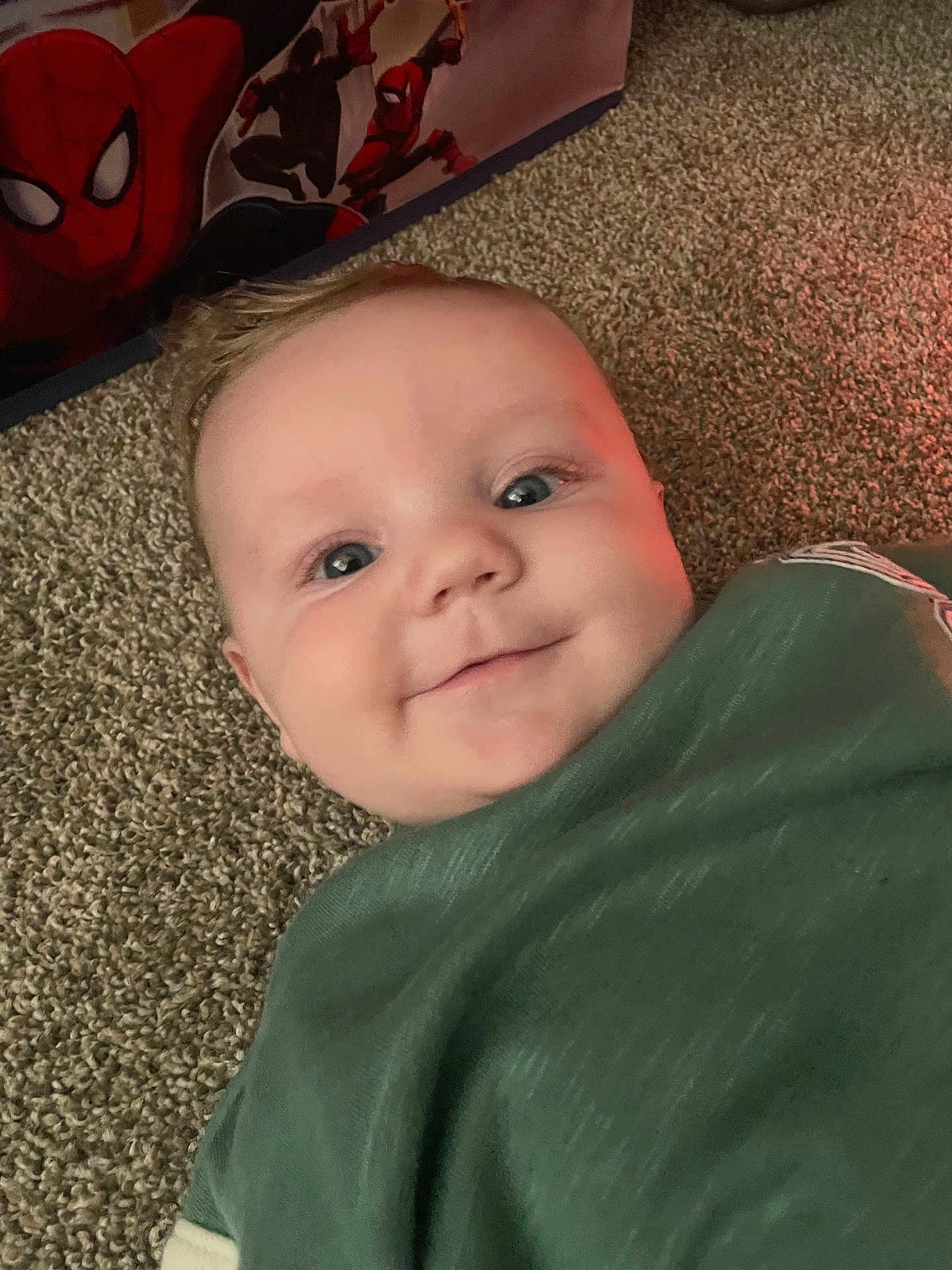 Atlas joined the competition — help win amazing prizes! baby, infant, smiling, carpet, blanket, green_clothing, face, eyes, cheeks, indoor, close_up, portrait, happy, lying_down, spiderman, superhero_poster, soft_light, cute, head, cozy