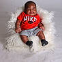 Emersyn is registered to the contest to win money with this photo: baby, infant, smiling, orange_shirt, nike_logo, gray_shorts, black_socks, fluffy_cushion, white_background, studio_photo, cute, happy, portrait, sitting, child, soft_texture, indoor, cozy, young_child, casual_clothing