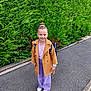 backpack, casual_clothing, child, coat, fashion, girl, greenery, hair_bun, happy, hedge, jacket, nature, outdoor, portrait, purple_pants, sidewalk, smile, standing, urban, white_shoes