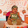 baby, child, high_chair, birthday, party, decorations, balloons, headband, smiling, happy, celebration, green_outfit, daisy, one, first_birthday, indoor, person, toddler, festive, colorful