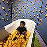 Jiquavious is registered to the contest to win money with this photo: bathtub, black_clothing, blue_wall, child, curly_hair, decor, floor, fun, green_carpet, indoor, playful, purple_carpet, room, rubber_duck, shadow, sitting, toddler, toy, wall, yellow