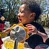 baby, black_clothing, child, colorful, curly_hair, daylight, hand, happy, infant, mickey_mouse, nature, outdoor, park, plastic_toy, playtime, profile, smiling, sunlight, toy, trees