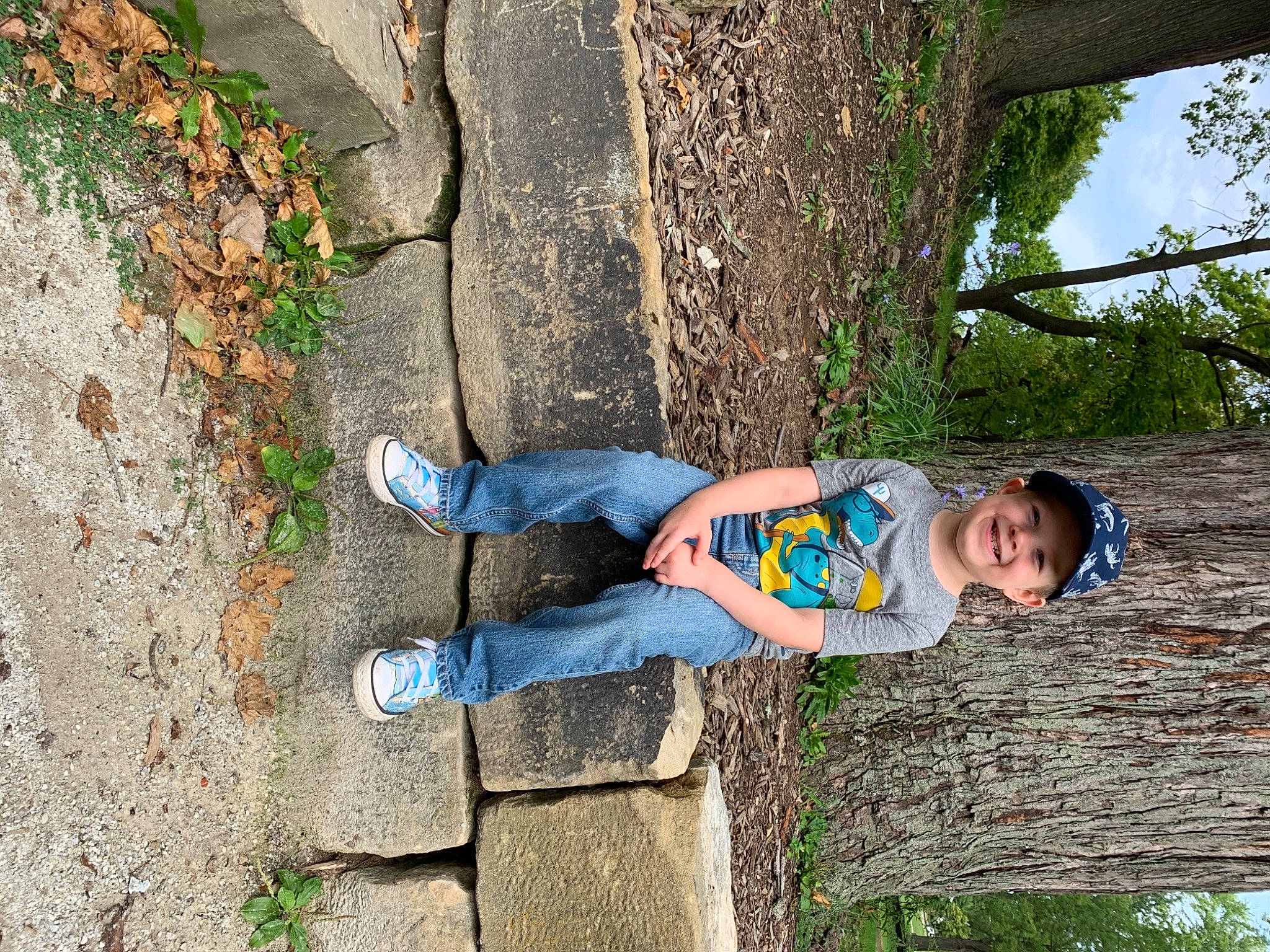 Kent is registered to the contest to win money with this photo: denim, electric_blue, fun, grass, happy, headwear, human_leg, joy, leg, leisure, people_in_nature, person, plant, recreation, sitting, smile, soil, toddler, tree, trunk