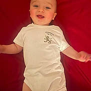 Alex participe au concours pour gagner de l'argent avec cette photo : baby, baby_clothing, cheeks, child, cute, eyes, face, happy, head, human, indoor, infant, lying_down, mouth, person, red_background, short_sleeves, skin, smiling, white_onesie
