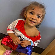 Arianna is registered to the contest to win money with this photo: bright_eyes, candid, casual, child, colorful_clothing, curly_hair, cute, face_tattoos, floor_tiles, happy, indoor, kneeling, pink_sneakers, playful, portrait, red_shirt, shorts, smiling, wall, young_child
