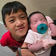 Jayzen is registered to the contest to win money with this photo: child, baby, newborn, pacifier, red_shirt, sleeping, holding, sibling, indoor, person, cute, face, happy, family, infant, love, skin, hair, hand, arm