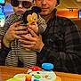 baby, child, young_man, indoor, plush_toy, mickey_mouse, sunglasses, striped_clothing, hat, table, toy, person, smile, holding, casual_clothing, wooden_table, face, headwear, playful, family