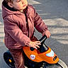 Nino participe au concours pour gagner de l'argent avec cette photo : child, corduroy_pants, curly_hair, expression, handlebars, hood, jacket, orange, outdoor, pavement, person, play, ride_on, shadow, small_wheels, sneakers, sticker, sunlight, toddler, toy_bike