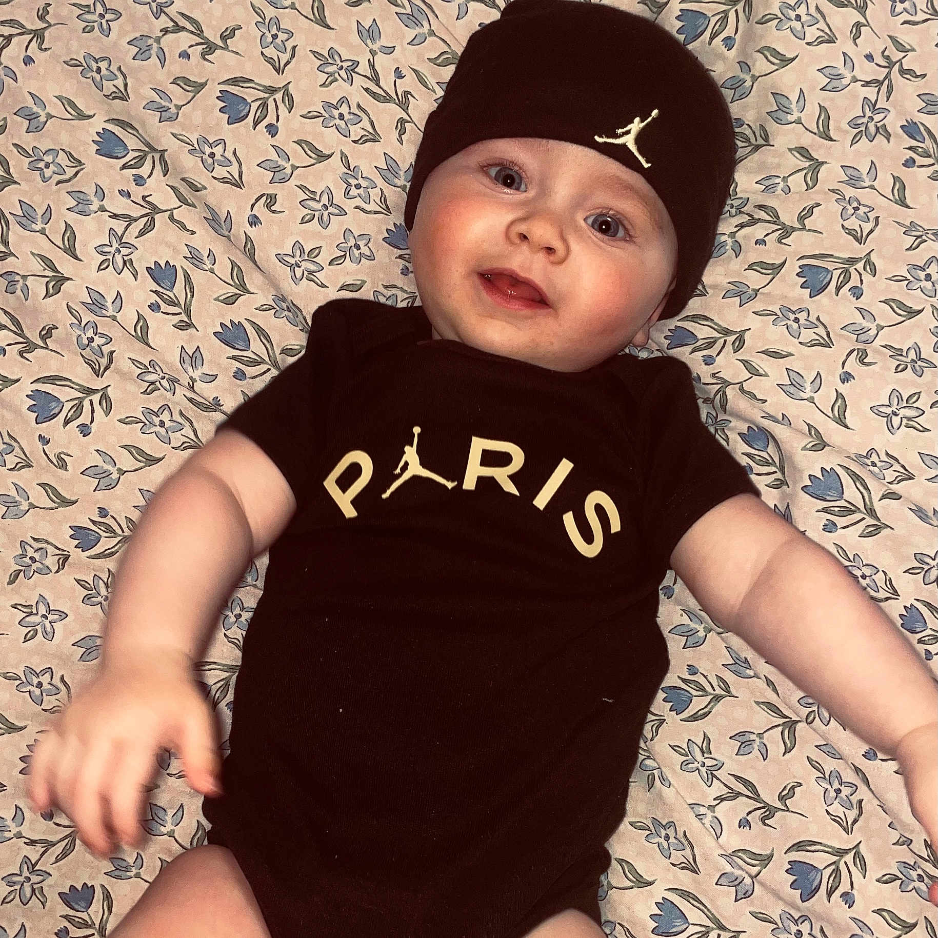 Sonny is registered to the contest to win money with this photo: arm, baby, baby_onesie, beanie, black_clothing, child, cute, eyes, face, floral_blanket, hand, headwear, indoors, infant, lying_down, mouth, person, portrait, skin, smiling