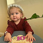 Victoria joined the competition — help win amazing prizes! baby, flower_crown, 10_months, wooden_sign, table, plant, indoor, child, curious, cute, portrait, clothing, long_sleeves, earrings, skin, hand, fingers, light, shadow, milestone