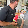 dog, man, couch, indoor, living_room, plant, toy, casual_clothing, happy, tongue_out, blue_eye, pet, home, seated, smiling_dog, shorts, black_tshirt, bracelet, vape, hanging_plant
